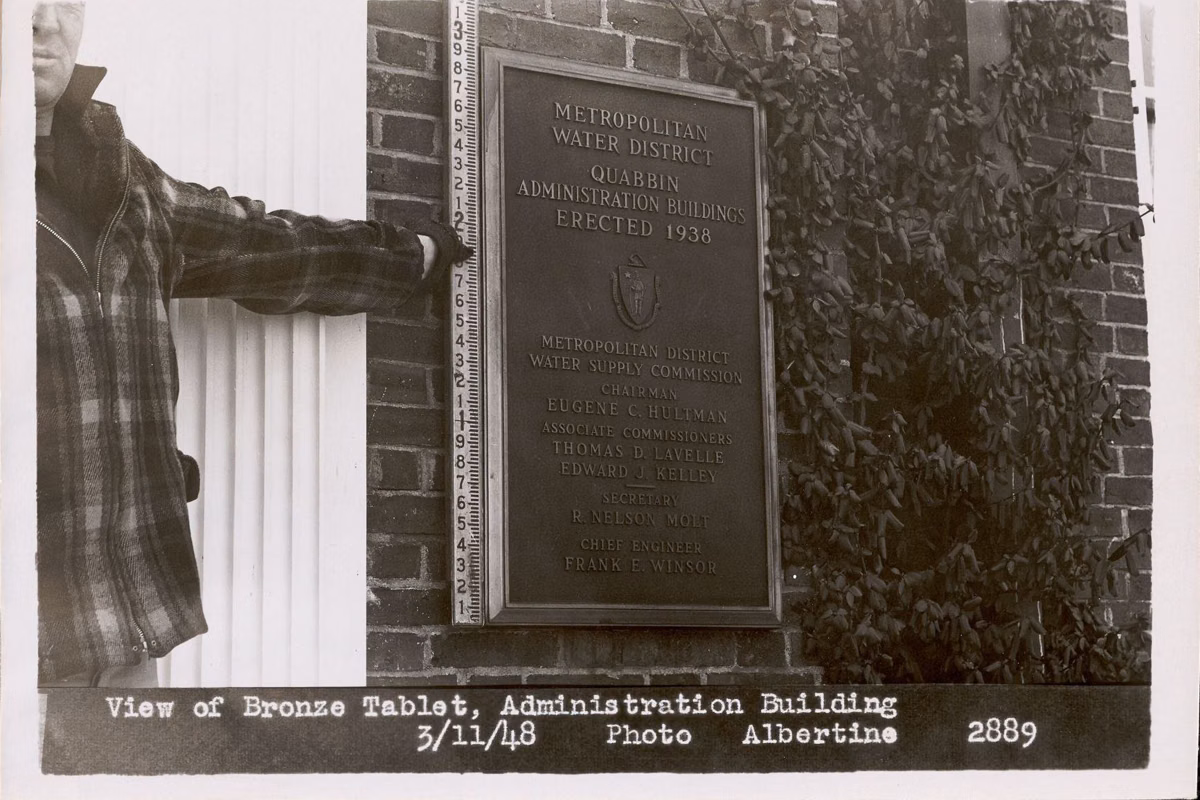 Black-and-white 1948 photo of a man beside a bronze tablet on the brick wall of the Quabbin Administration Building